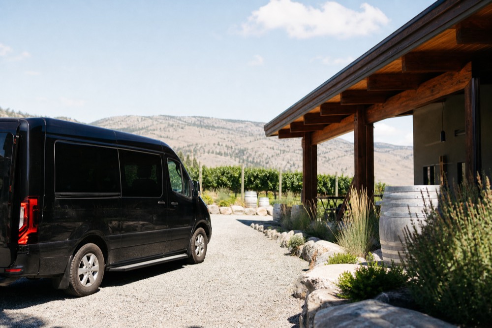 A small-group tour van parked outside a boutique winery tasting room surrounded by vines on the Naramata Bench, midday sun, no people, no plate, no branding.