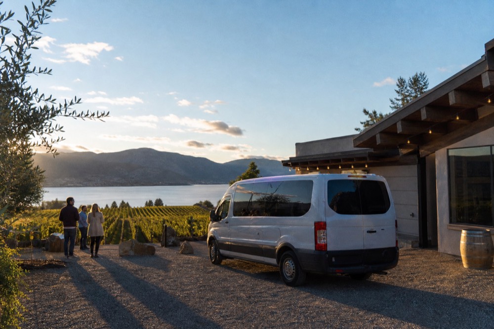 A small tour van parked beside a vineyard tasting room under blue sky, no branding, no plate.