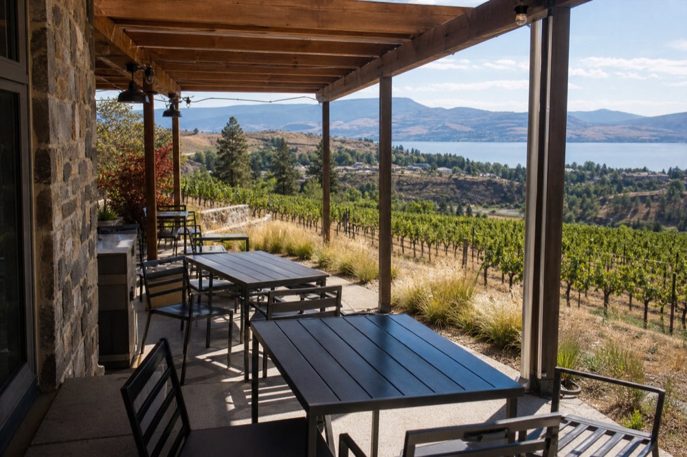 A quiet boutique cellar-door tasting patio on an East Kelowna slope, wooden pergola over a few tables, vineyard rows dropping toward the lake, warm morning light, no people, no logos.