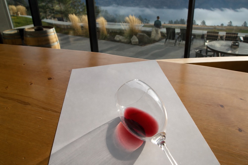 Overhead view of a white tasting sheet with a wine glass tilted to show colour against the white paper, on a sunlit tasting-room table, no people, no text.