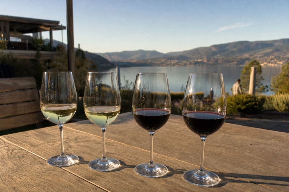 A tasting flight of red and white wine glasses on an outdoor patio table overlooking Okanagan Lake at golden hour, no people, no logos.