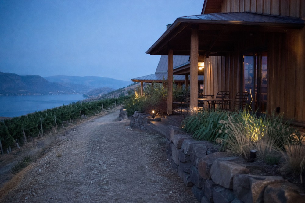 Cluster of small boutique winery tasting rooms along a narrow bench road with vineyard rows and a lake view below, late afternoon light, no people, no signage.