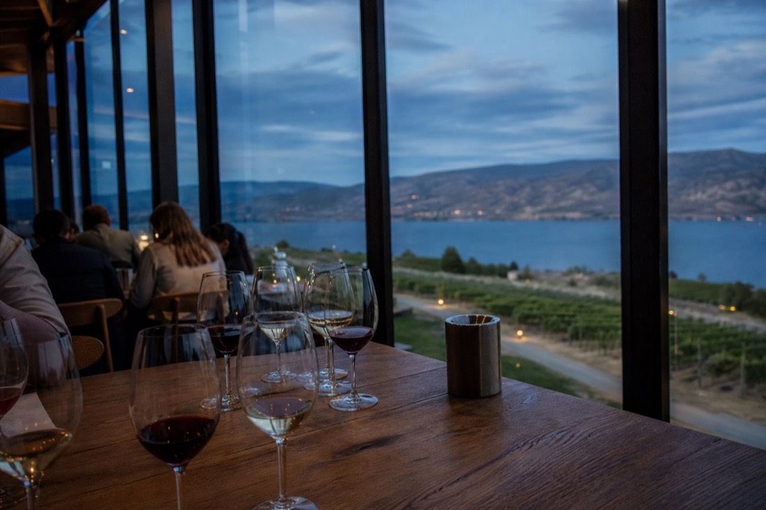 A cellar-door tasting bench with several wine glasses poured for a small group, vineyard rows and a big blue lake visible through tall windows in warm light