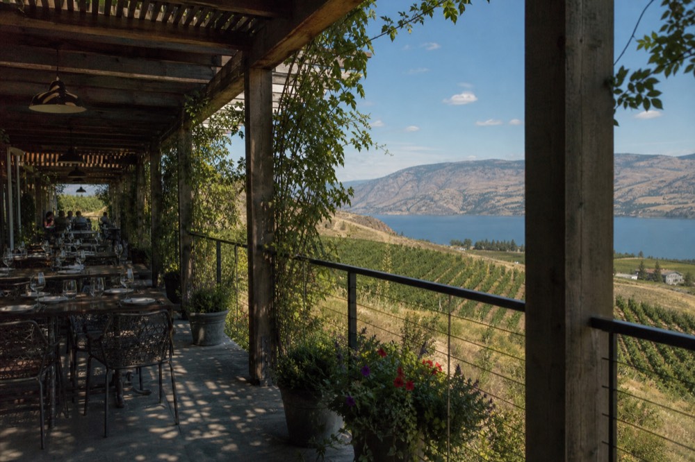 A long pergola-shaded winery terrace with tables set for lunch, vines below and a blue lake beyond under bright sky, no people, no logos, no text.