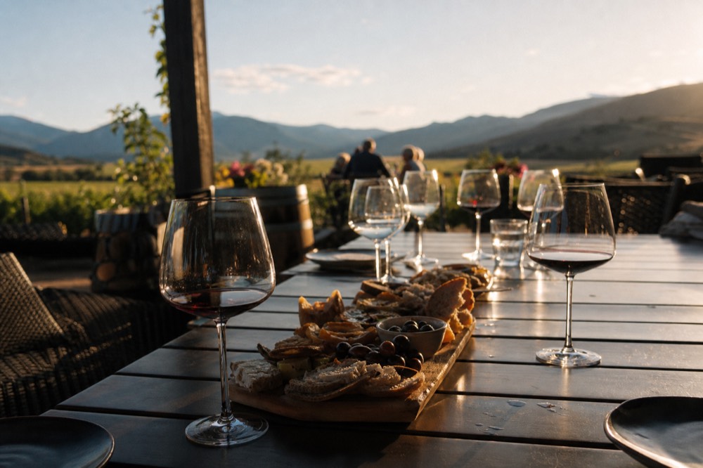 A row of wine glasses and a shared platter of food on a long outdoor table at a winery at golden hour, warm low light across the vines, no people, no logos, no text.
