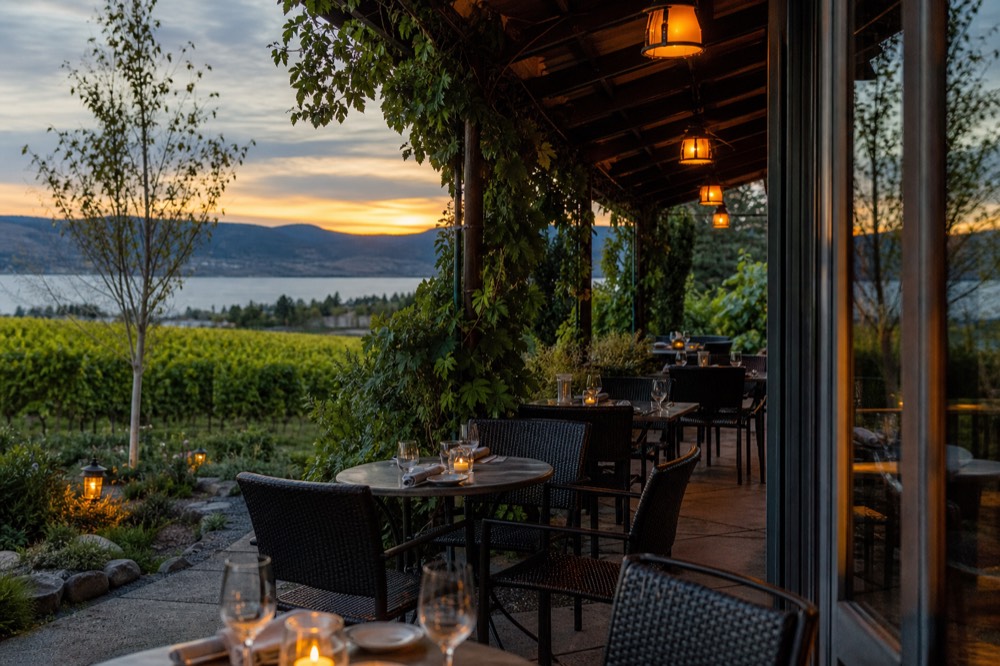 An evening winery patio with lamps lit and tables set for dinner as the sun sets over a calm lake behind the vines, warm amber light, no people, no branding, no text.