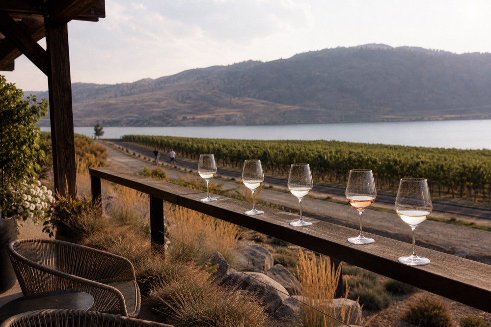 A sparkling-wine tasting room patio on the lower bench overlooking the lake and the old rail trail, glasses on a rail, no people, no branding, no text.