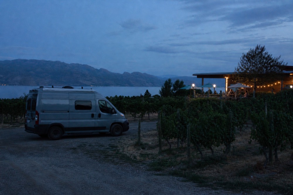 A small touring van parked at the head of a vineyard row with a tasting patio beyond, lake in the distance, no people, no logos, no text.
