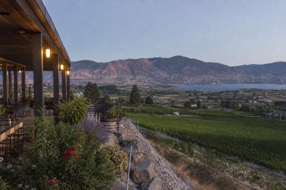 A hillside winery patio looking out over a broad valley of vineyard blocks toward dry mountains and a distant lake at Oliver, warm afternoon light, no people, no branding, no text.