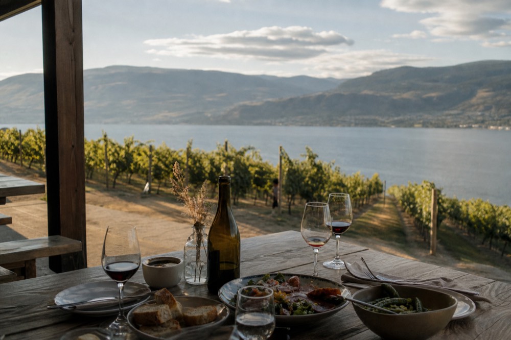 A rustic outdoor table set with wine glasses and a light lunch spread on a bench-top vineyard patio with the lake below, soft midday light, no people, no branding, no text.