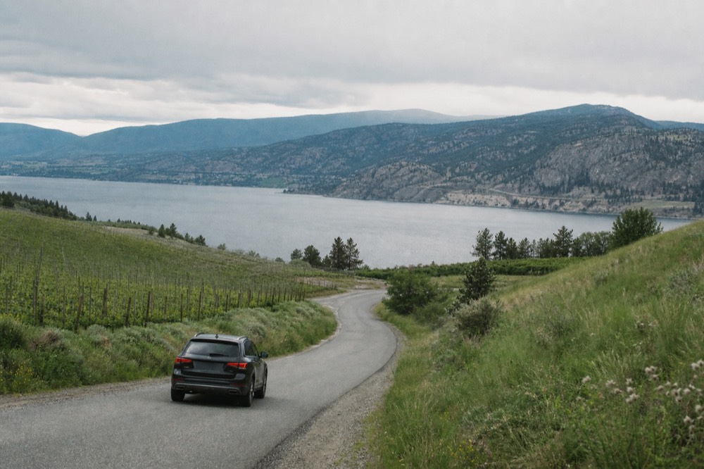 A single vehicle on an open vineyard road winding through hills toward Okanagan Lake, no branding, no people, no signage.
