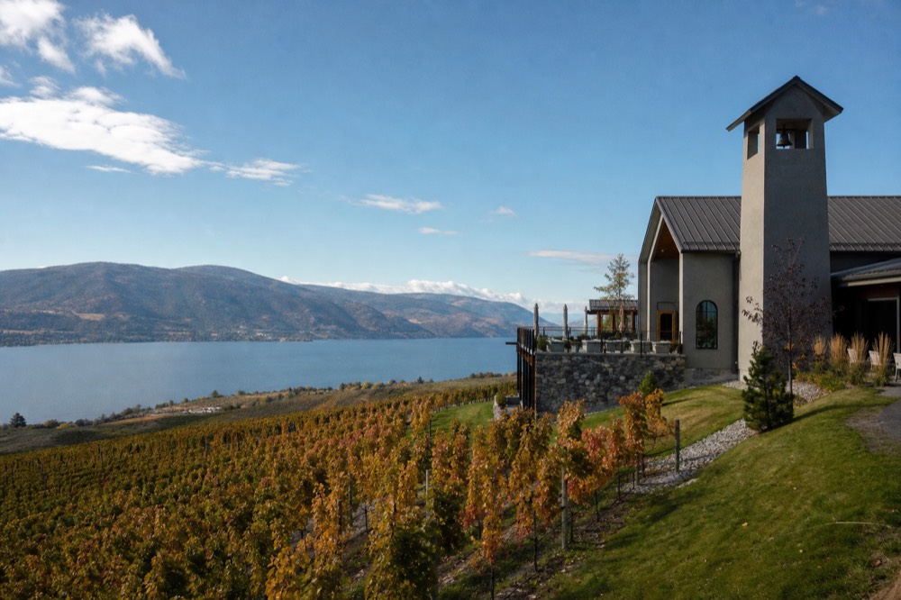 A modern hilltop winery building with a tall bell tower and stone terrace overlooking neat vineyard rows and a wide blue lake, no people, no logos, no text.